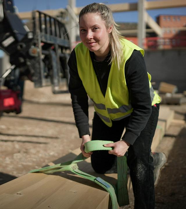 Eine Frau in gelber Sicherheitsweste beugt sich über einen langen Holzbalken und hat einen Gurt in der Hand. Im Hintergrund ist eine Baustelle zu sehen.