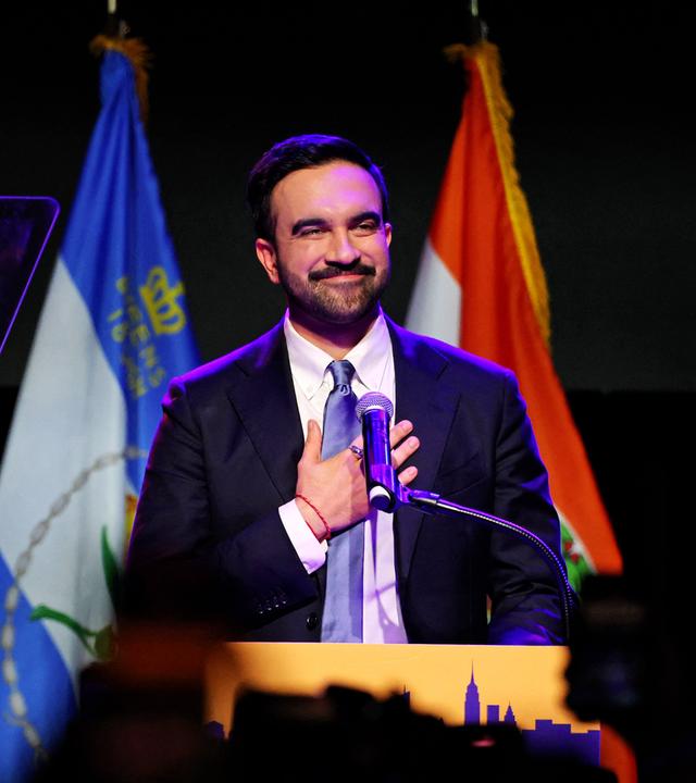 Ein Mann mit Bart und im Anzug steht hinter einem Rednerpult vor Flaggen und legt sich lächelnd eine Hand aufs Herz