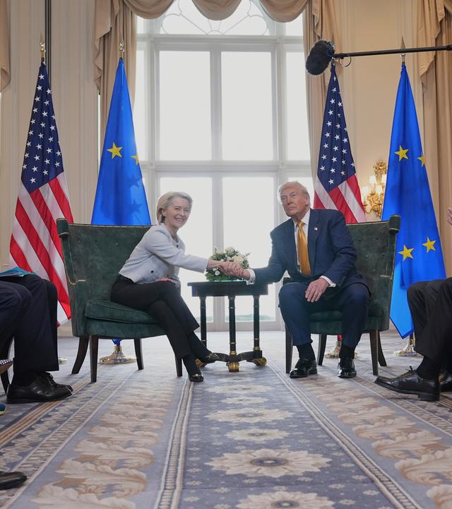 Ursula von der Leyen und Donald Trump geben sich zum Deal die Hände. Hinter ihnen stehen EU- und US-Flaggen