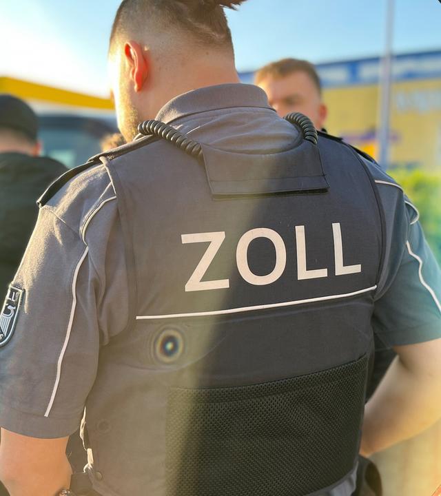 Eine Gruppe Zollbeamten und Zollbeamtinnen steht im Freien. Zentral steht ein Beamter mit dem Rücken zur Kamera. Seine Stichschutzweste trägt den Schriftzug "Zoll".