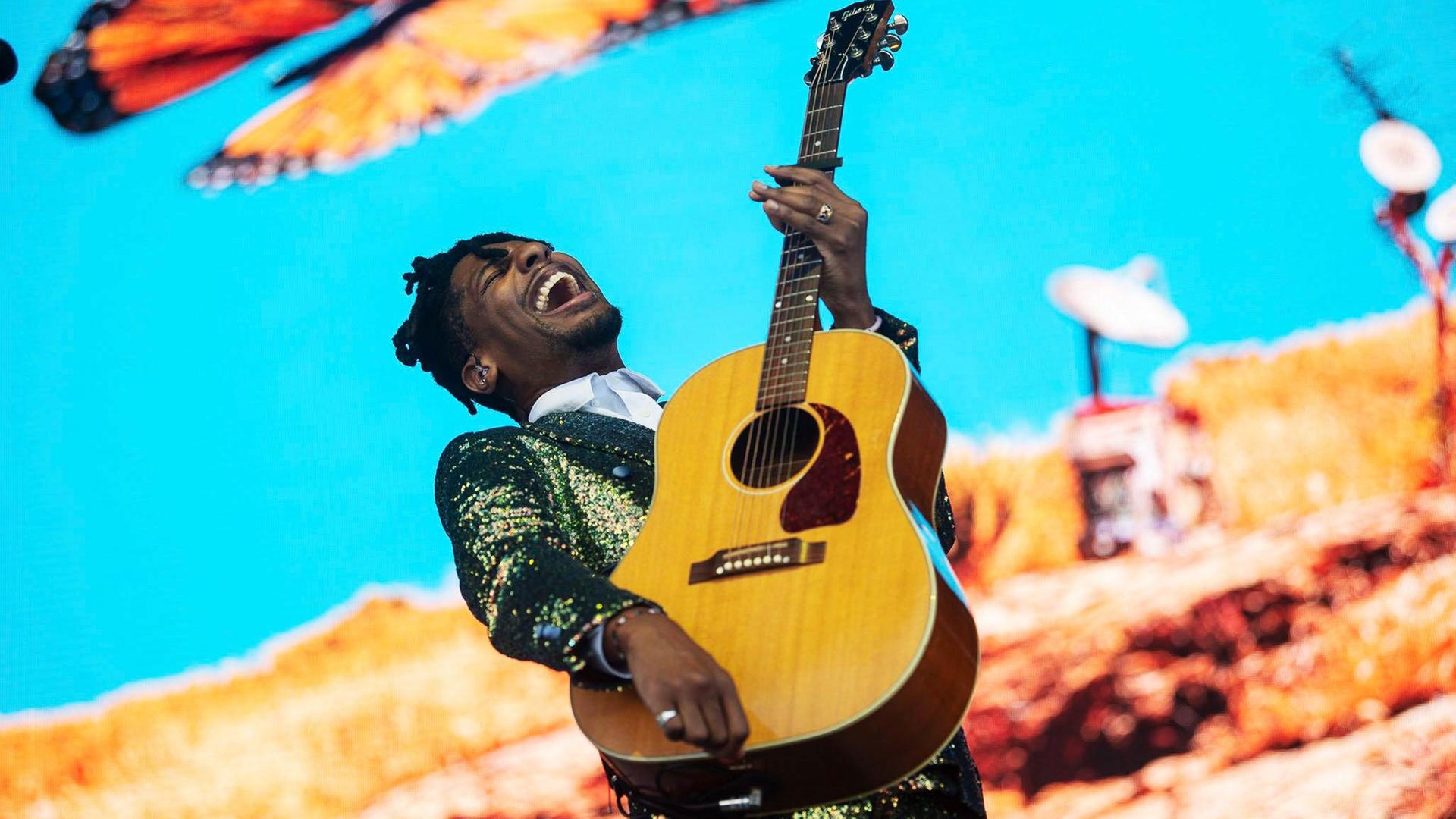 Ein Mann mit Gitarre in der Hand singt vor hellblauem Hintergrund. Links über ihm sieht man einen großen Schmetterling.