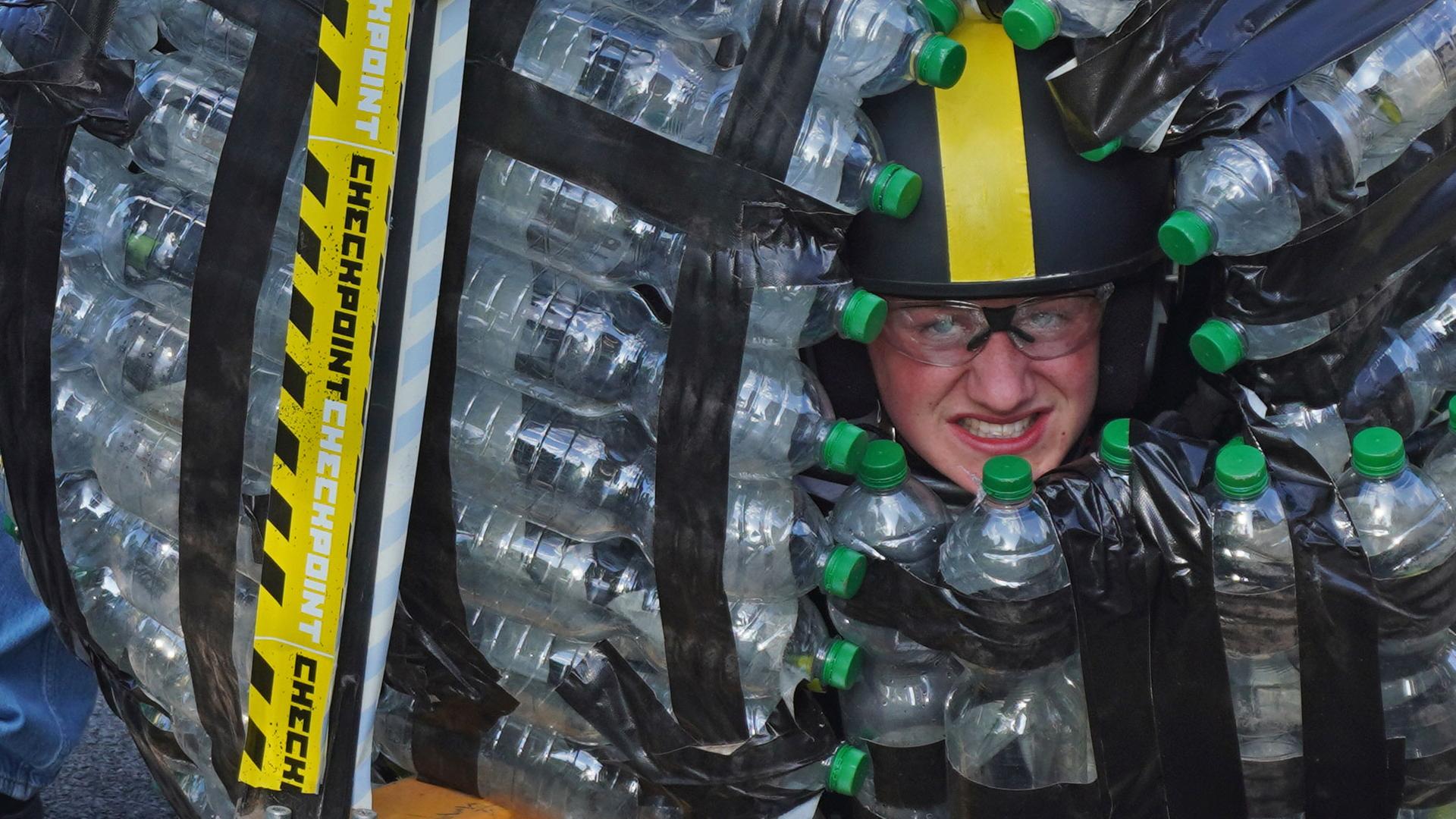 Checkpoint: Vencent sitzt in einem Zorbingball aus Pfandflaschen, aus dem nur sein behelmter Kopf herausschaut. Die anderen zwei Moderatoren sitzen um ihn herum und grinsen herausfordernd in die Kamera.