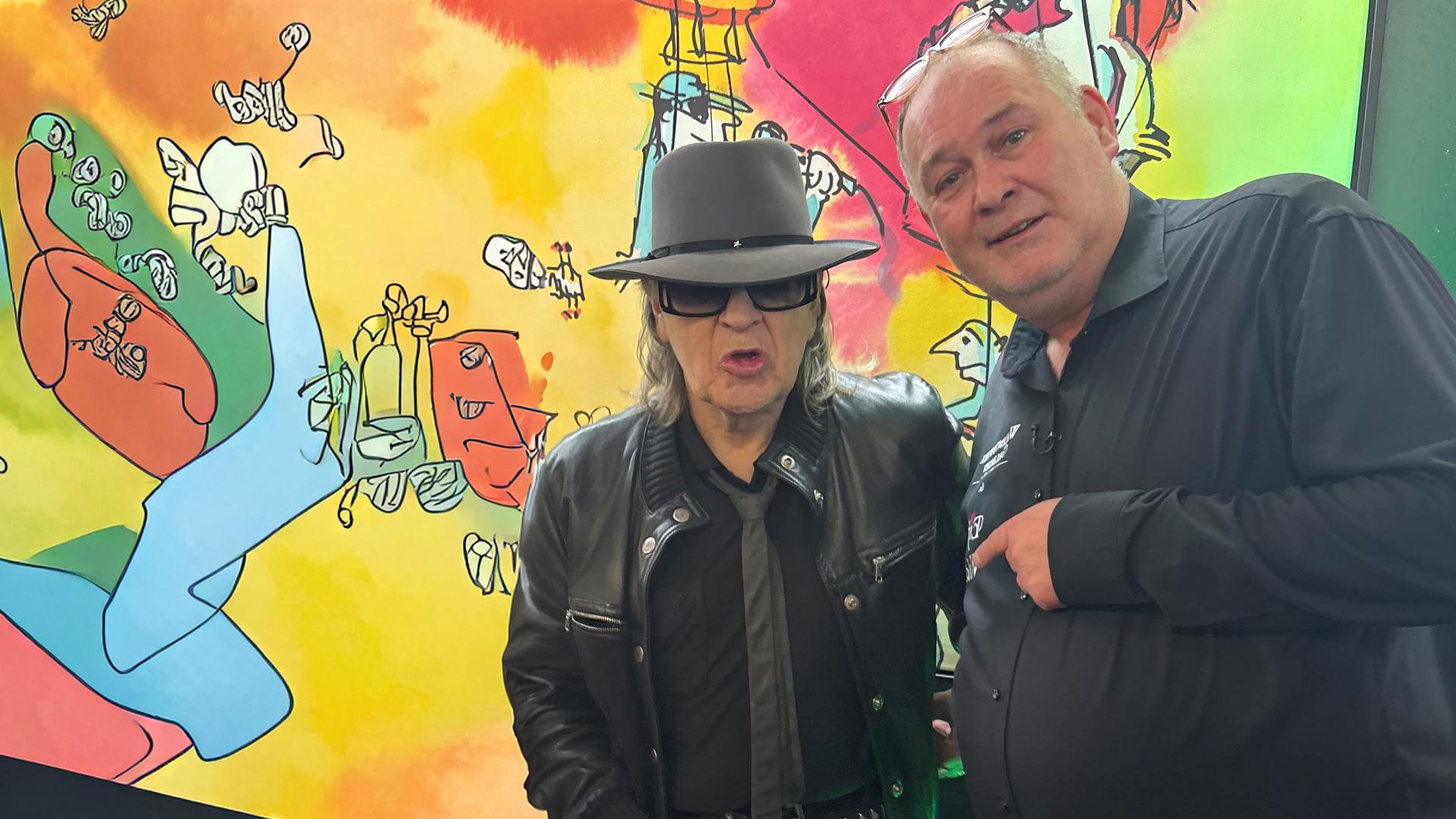Udo Lindenberg und Waldi stehen vor einem bunten Gemälde in einer Galerie und schauen beide in die Kamera.