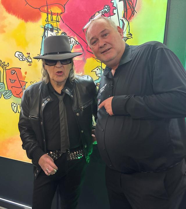 Udo Lindenberg und Waldi stehen vor einem bunten Gemälde in einer Galerie und schauen beide in die Kamera.
