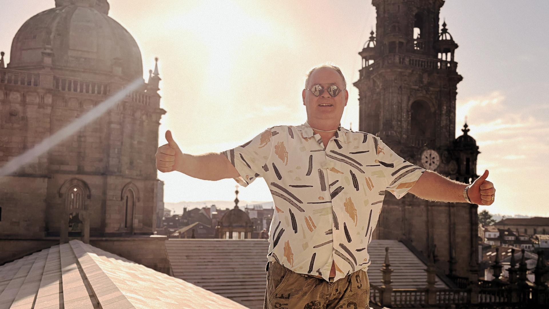 Waldi steht mit Sonnenbrille und ausgebreiteten Armen vor einem sonnigen Himmel. Im Hintergrund der Glockenturm einer Kirche.