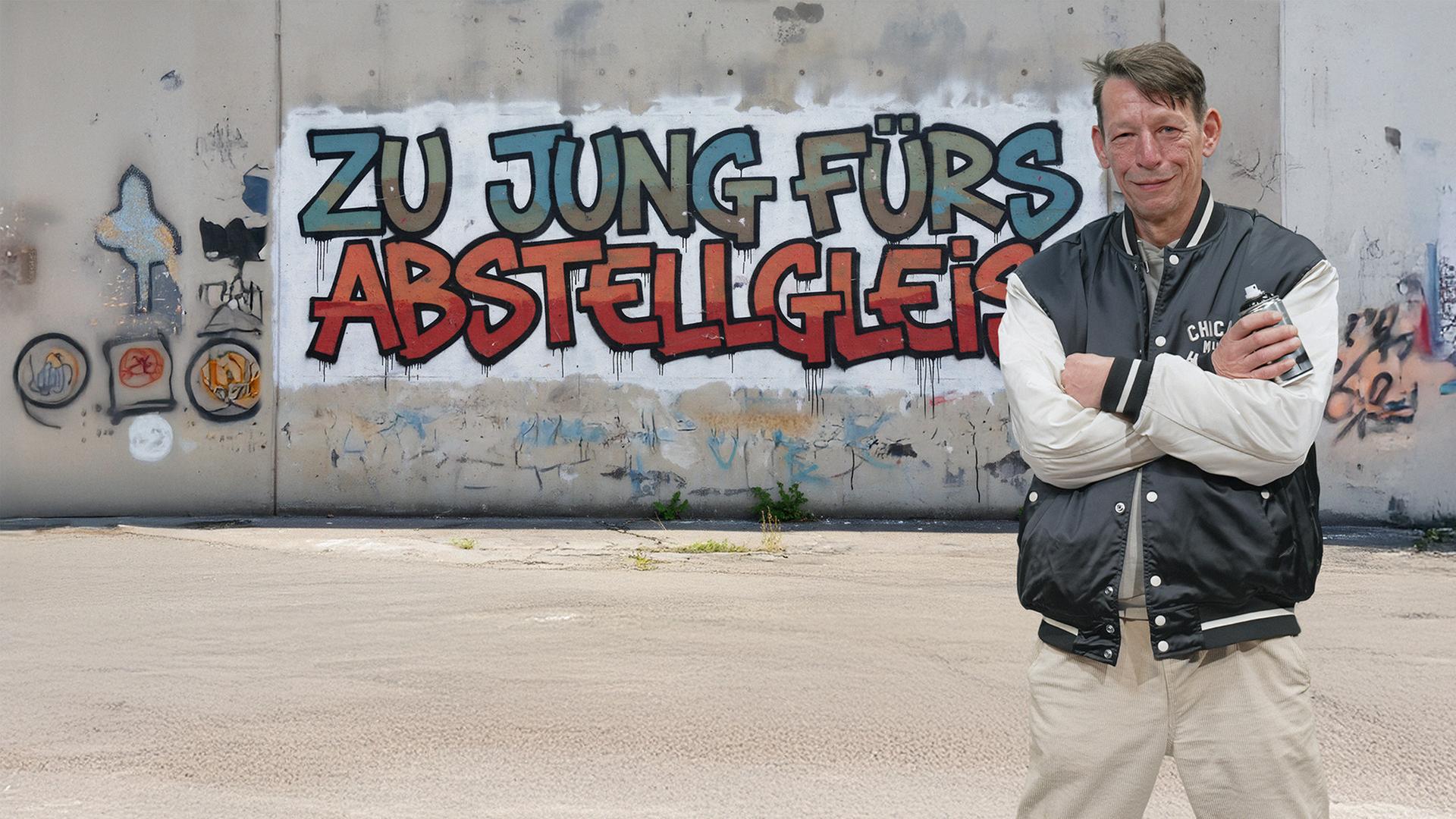Klemens Schüttken (58)  steht vor einem Graffiti an einer Betonwand. Er hat die Arme vor der Brust verschränkt und schaut in die Kamera. Auf der Wand steht geschrieben "ZU JUNG FÜRS ABSTELLGLEIS".