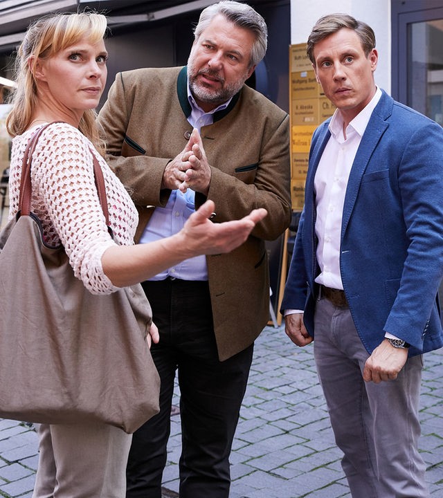 "Die Rosenheim-Cops - Zu Tode gekuschelt": Sabine Stadelmayer (Jenny-Marie Muck) steht den Kommissaren Stadler (Dieter Fischer) und Bach (Patrick Kalupa) in einer Fußgängerzone gegenüber. Kommissar Stadler sieht sie an, Sabine Stadelmayer schaut nach rechts. Sie trägt eine große braune Tasche über der Schulter.