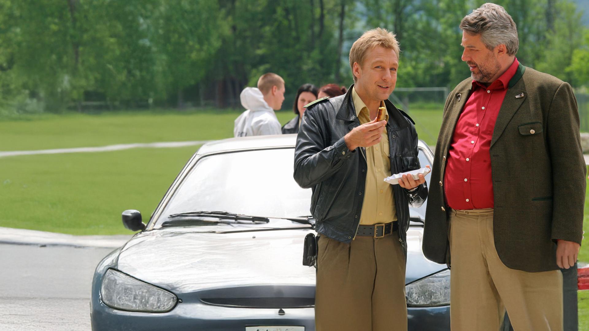 "Die Rosenheim-Cops - Zu Tode gesteigert:" Am Tatort stehen Polizist Mohr (Max Müller) und Kommissar Stadler (Dieter Fischer) an einem Auto, und reden miteinander.