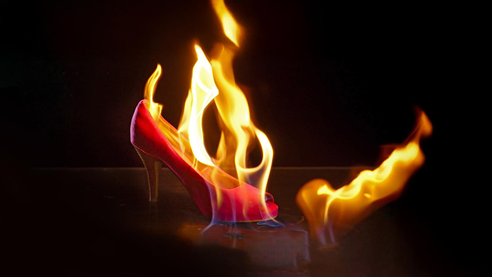 Im Zentrum des Bildes steht ein brennender pinker Stöckelschuh. Der Hintergrund ist schwarz. 