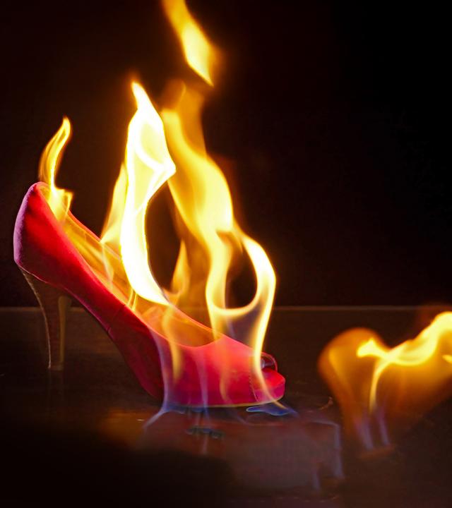 Im Zentrum des Bildes steht ein brennender pinker Stöckelschuh. Der Hintergrund ist schwarz. 