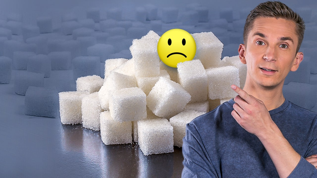 Eric Mayer im Vordergrund, dahinter jede Menge Würfelzucker. Eric zeigt mit der linken Hand auf einen kleinen Berg gestapelt aus Würfelzucker, auf dem ein negativer Smiley sitzt.