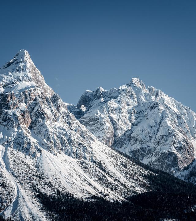Das Bild zeigt eine beeindruckende Winterlandschaft in den Tiroler Alpen, speziell in der Zugspitzregion. Im Vordergrund sind schneebedeckte Berge sichtbar, deren Gipfel in verschiedenen Höhen aufragen. Der markanteste Berg ist spitz und von einer geschwungenen Schneeschicht bedeckt.   Im unteren Bereich des Bildes sind bewaldete Hänge zu sehen, die ebenfalls mit Schnee bedeckt sind. Der Himmel ist klar und strahlend blau, was einen starken Kontrast zur weißen Schneelandschaft bietet.   Das Bild vermittelt eine kalte, klare Winternatur, die die majestätische Schönheit der Alpenlandschaft zur Geltung bringt.