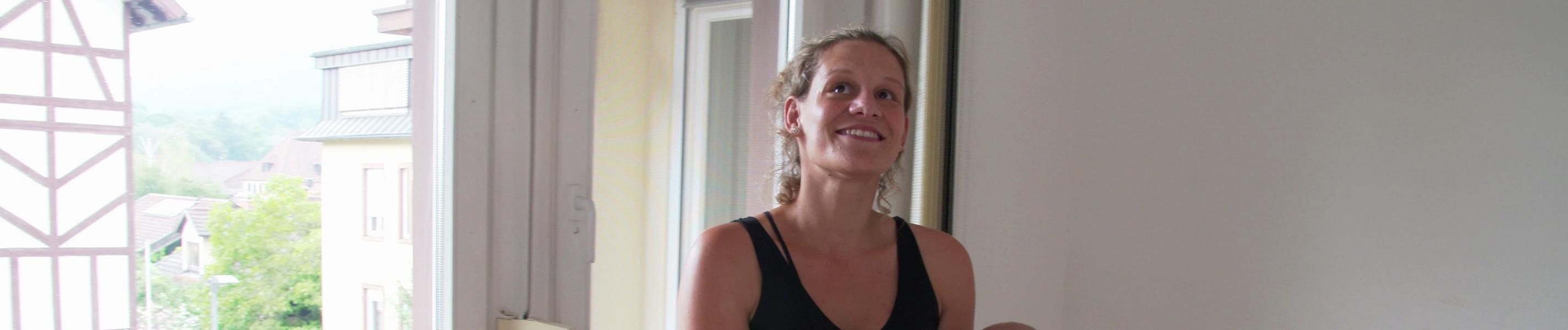 Eine junge Frau in einem leeren Raum vor der Balkontür stehend mit einem schlafenden Baby auf dem Arm. Sie lacht, wirkt hoffnungsvoll.