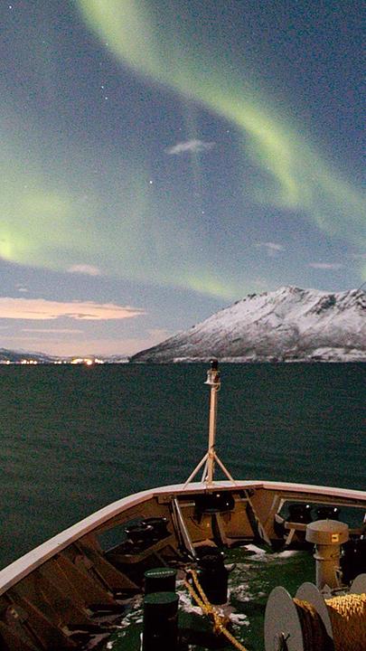Polarlichter an einem winterlichen Nachmittagshimmel. Im Vordergrund ist die Spitze eines Schiffs erkennbar