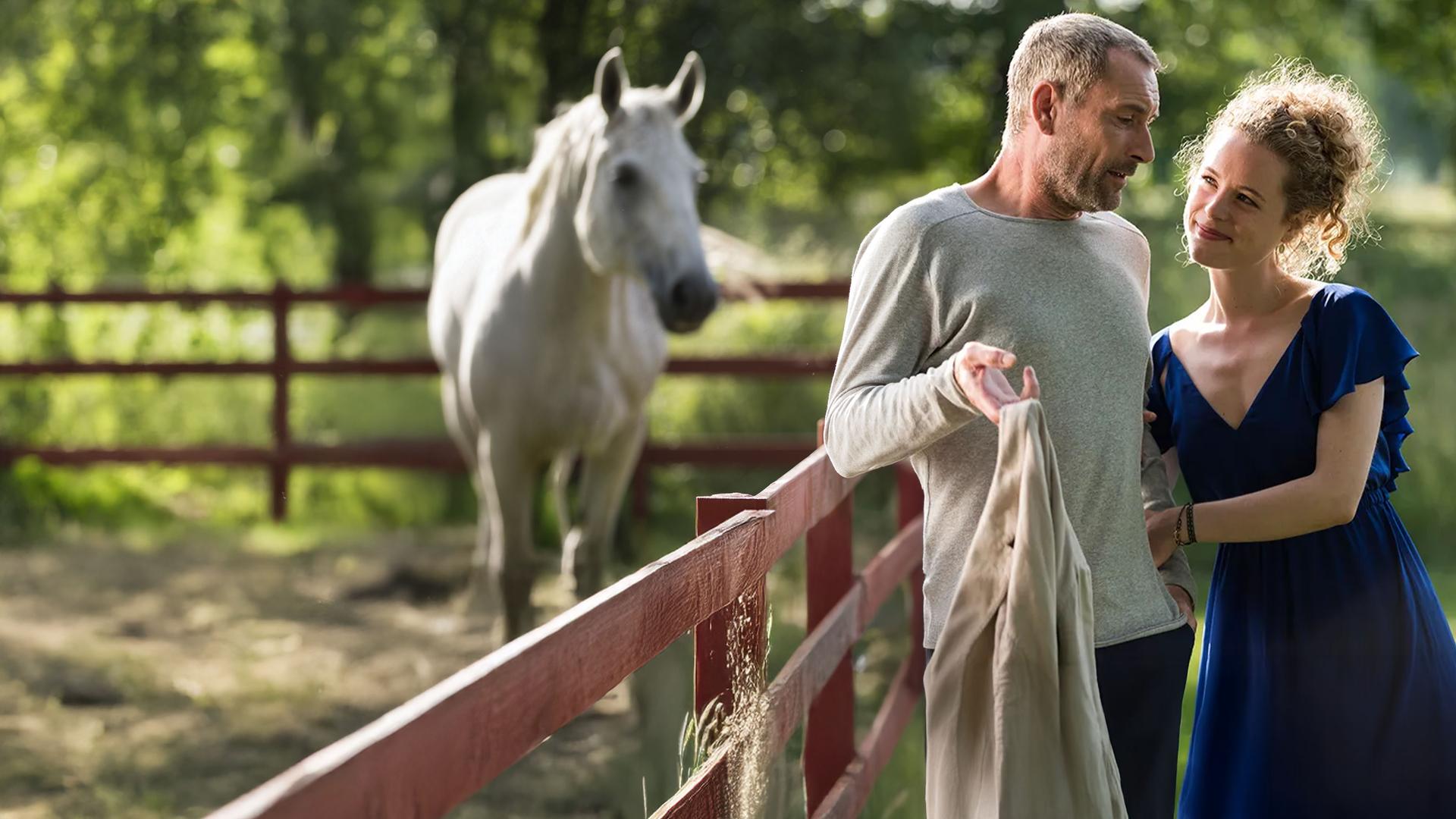 Emilia Jönsson (Klara Deutschmann) hängt im Arm von Caspar Jönsson (Heikko Deutschmann), der an einem Zaun lehnt. Im Hintergrund steht ein weißes Pferd.