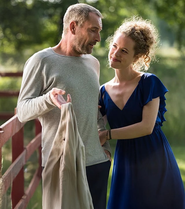 Emilia Jönsson (Klara Deutschmann) hängt im Arm von Caspar Jönsson (Heikko Deutschmann), der an einem Zaun lehnt. Im Hintergrund steht ein weißes Pferd.