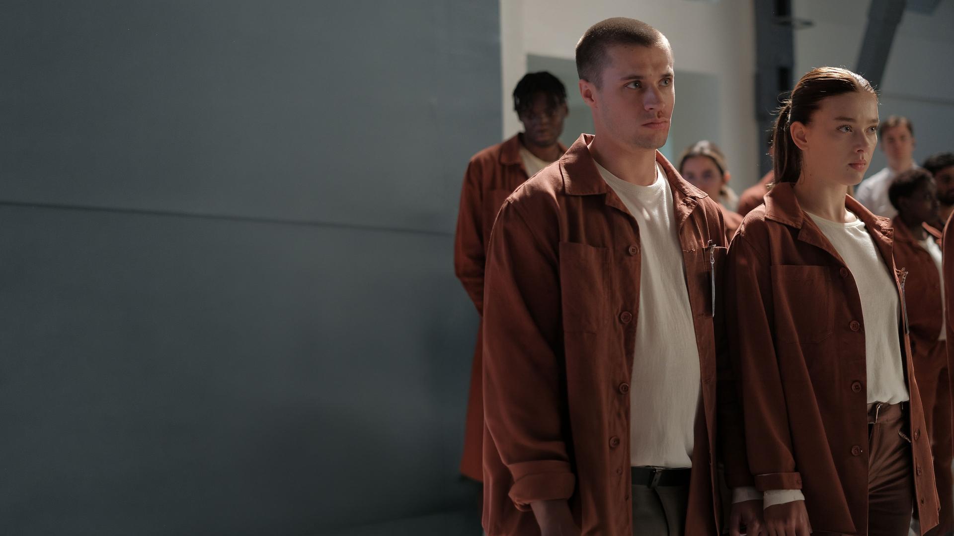 "Fallen - Zusammenhalt": Luce (Jessica Alexander) und Daniel (Gijs Blom) stehen nebeneinander in einer Gruppe von Personen (Komparsen), alle tragen orangene Anzüge mit Namensschildern. Beide blicken ernst geradeaus. Im Hintergrund sind karge, weiße Wände mit einem Stahlpfeiler zu erkennen.