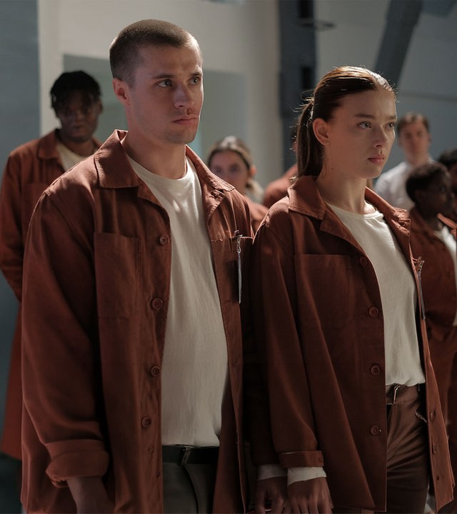 "Fallen - Zusammenhalt": Luce (Jessica Alexander) und Daniel (Gijs Blom) stehen nebeneinander in einer Gruppe von Personen (Komparsen), alle tragen orangene Anzüge mit Namensschildern. Beide blicken ernst geradeaus. Im Hintergrund sind karge, weiße Wände mit einem Stahlpfeiler zu erkennen.