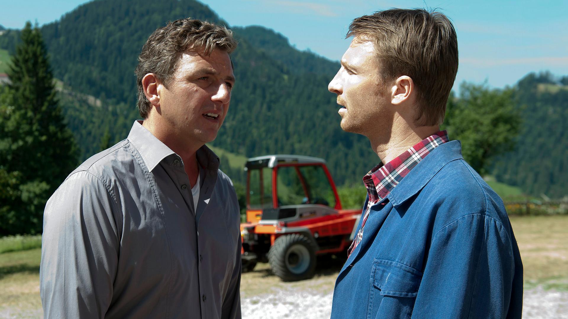 "Der Bergdoktor - Zwänge": Martin (Hans Sigl) und Hans (Heiko Ruprecht) stehen auf einem Feld und unterhalten sich. Im Hintergrund ein Traktor und das Bergpanorama.