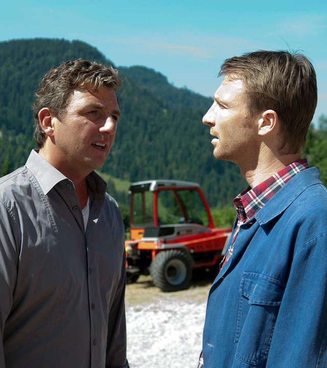 "Der Bergdoktor - Zwänge": Martin (Hans Sigl) und Hans (Heiko Ruprecht) stehen auf einem Feld und unterhalten sich. Im Hintergrund ein Traktor und das Bergpanorama.