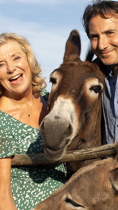 Jutta Riedl (Jutta Speidel) und Bruno Bertani (Bruno Maccallini) stehen an einem Zaun umringt von Eseln und lachen. Jutta füttert einen Esel mit einer Karotte.