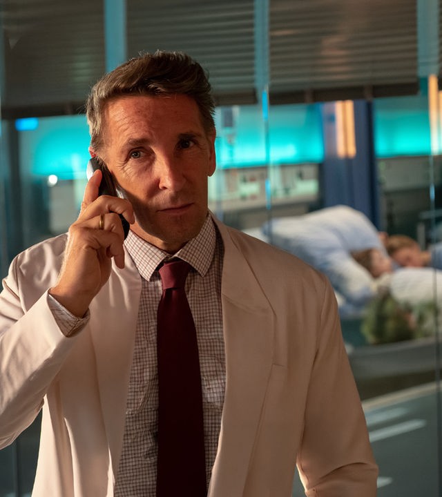 Vor einer Glasscheibe steht Dr. Kahnweiler (Mark Keller) und telefoniert mit seinem Handy. Hinter ihm im Zimmer liegt ein kleiner Junge im Krankenbett. Eine Frau hat ihren Oberkörper neben ihm abgelegt.