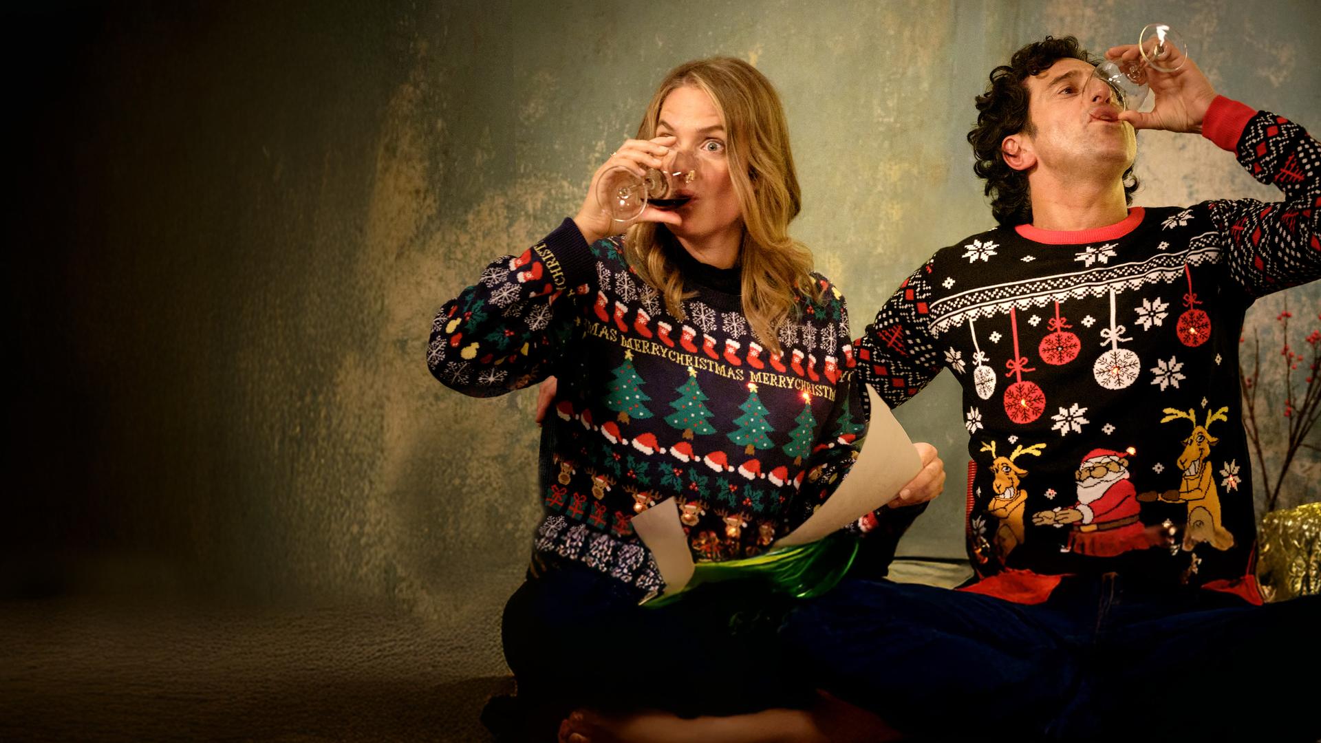 "Zwei Weihnachtsmänner sind einer zu viel": Karo und Lasse sitzen an einem Tisch und tragen bunte Pullover mit weihnachtlichen Motiven und LED-Lichtlein. Karo hält ein Blatt in der Hand. Beide nehmen einen kräftigen Schluck aus ihren Rotweingläsern.