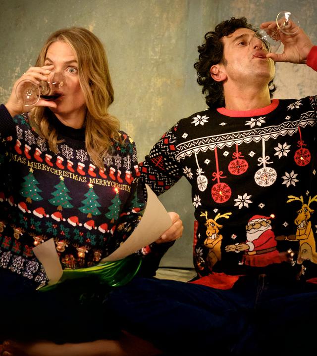 "Zwei Weihnachtsmänner sind einer zu viel": Karo und Lasse sitzen an einem Tisch und tragen bunte Pullover mit weihnachtlichen Motiven und LED-Lichtlein. Karo hält ein Blatt in der Hand. Beide nehmen einen kräftigen Schluck aus ihren Rotweingläsern.