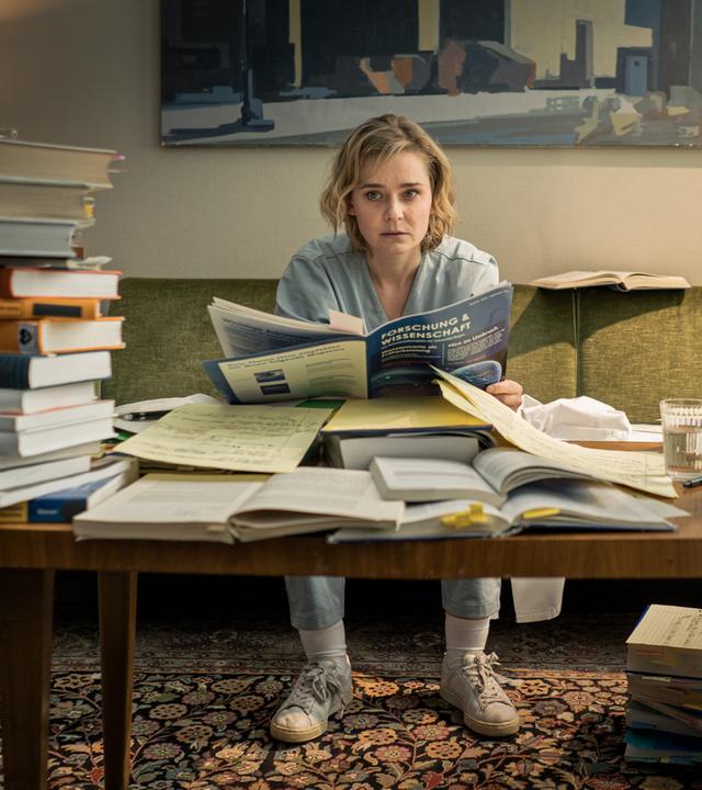 "Doktor Ballouz - Zweite Chance": Dr. Michelle Schwan (Nadja Bobyleva) sitzt vor Unmengen an gestapelten medizinischen Büchern an einem Tisch auf dem Sofa.