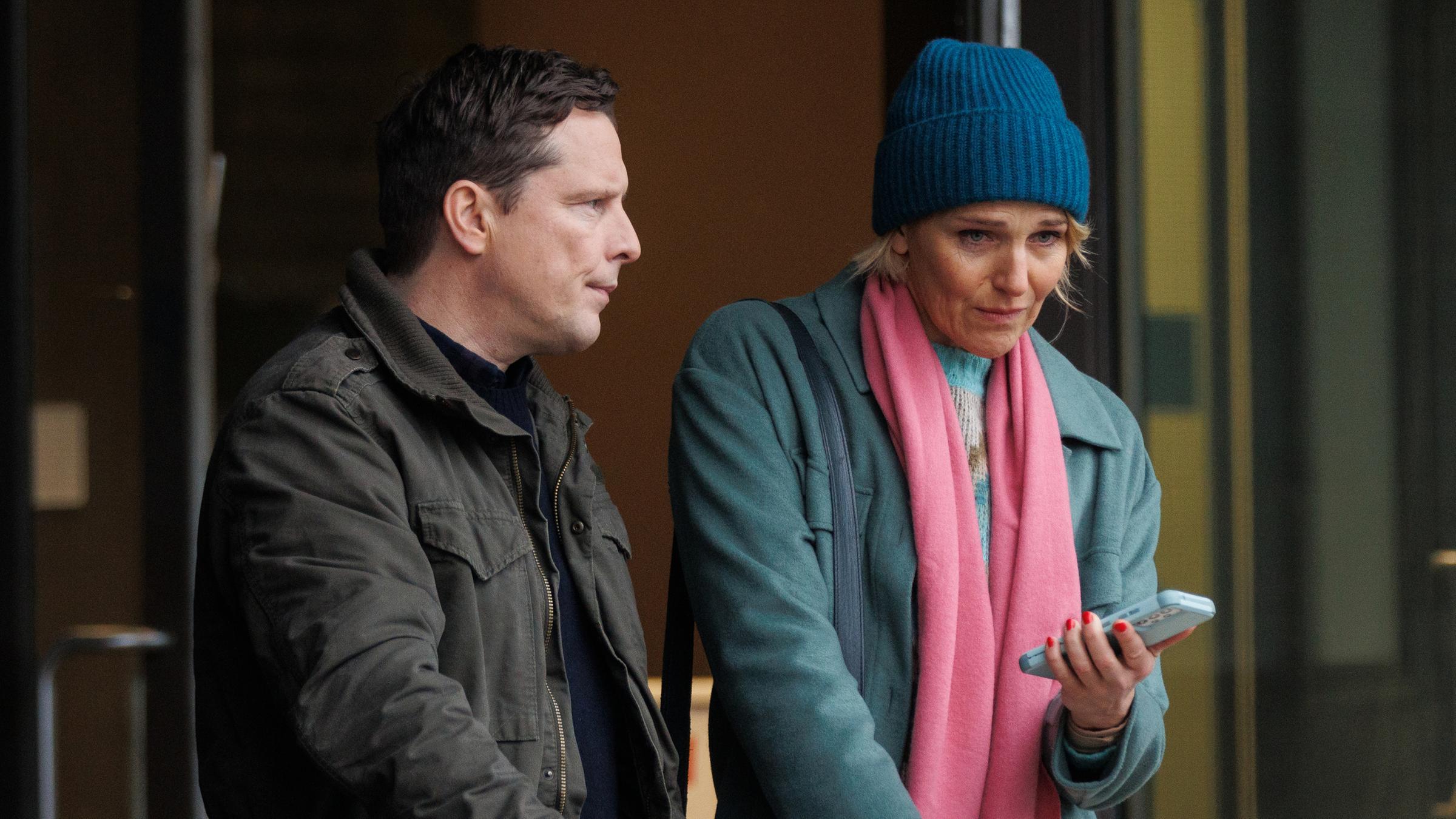 Fritzie Kühne (Tanja Wedhorn) hält ihr Smartphone in der Hand. Neben ihr steht Stefan Kühne (Florian Panzner). Beide machen ein sehr ernstes Gesicht.