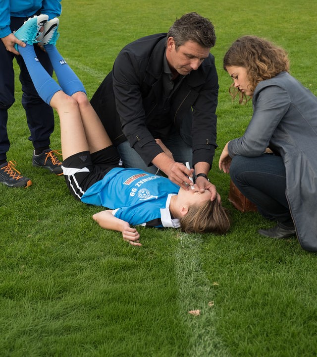 "Der Bergdoktor: Zwiespalt(1)": Martin Gruber (Hans Sigl) untersucht Fußballspielerin Paula Gaubinger (Leonie Wesselow), die verletzt am Boden liegt. Lilli Gruber (Ronja Forcher) hilft ihm dabei.