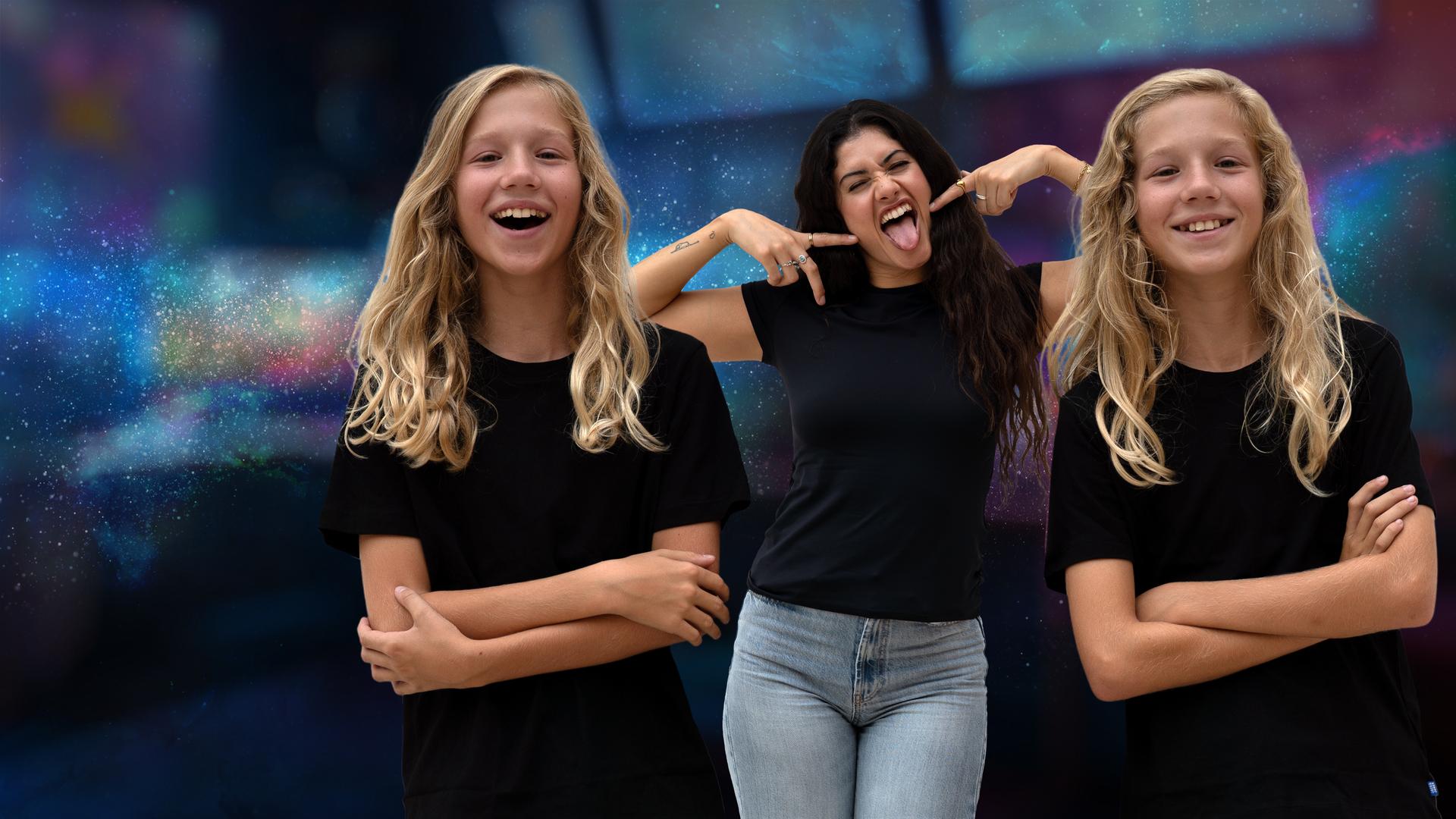 Die eineiigen Zwillinge Vin und Lex lächeln mit verschränkten Armen in die Kamera. Zwischen ihnen steht Jeannie Wagner und streckt die Zunge raus. Gemeinsam stehen sie vor einem bunten Hintergrund.