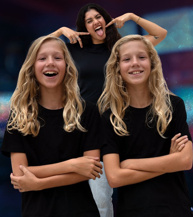 Die eineiigen Zwillinge Vin und Lex lächeln mit verschränkten Armen in die Kamera. Zwischen ihnen steht Jeannie Wagner und streckt die Zunge raus. Gemeinsam stehen sie vor einem bunten Hintergrund.