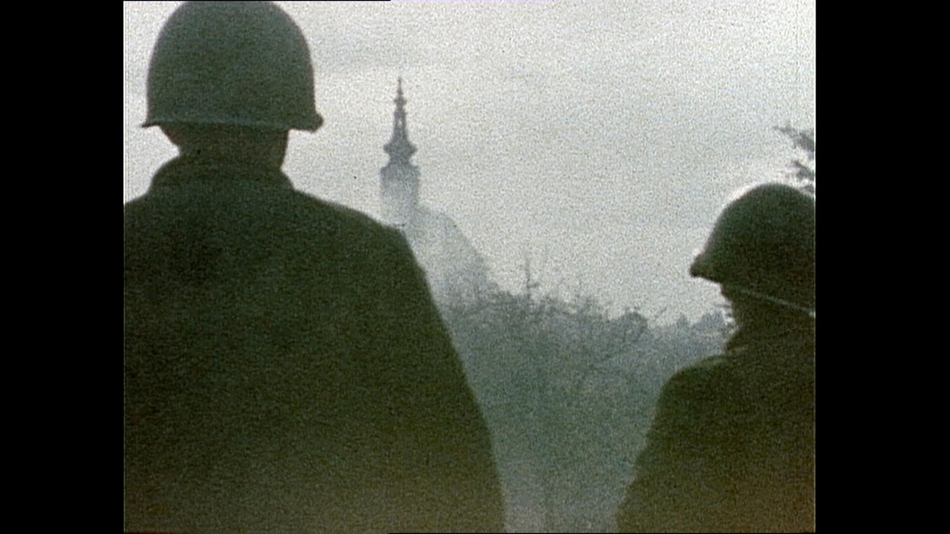 Das Bild zeigt zwei amerikanische Soldaten im Jahr 1945, die in Richtung einer unscharfen Stadtlandschaft blicken. Beide Soldaten tragen helle Stahlhelme und sind von hinten sichtbar. Sie stehen zusammen und blicken auf einen im Hintergrund leicht erkennbaren Turm, der Teil eines Gebäudes ist. Der Himmel über ihnen ist durch einen grauen Schleier verhangen, und die gesamte Szene hat eine melancholische und diffuse Lichtstimmung. Die Umgebung wirkt düster und lässt den Eindruck von Unruhe und Unsicherheit in der Luft entstehen. Es ist klar, dass es sich bei der dargestellten Situation um ein historisches Ereignis zur Zeit des Kriegsendes handelt, wobei die Kulisse vermutlich eine österreichische Stadt im Frühjahr 1945 darstellt.