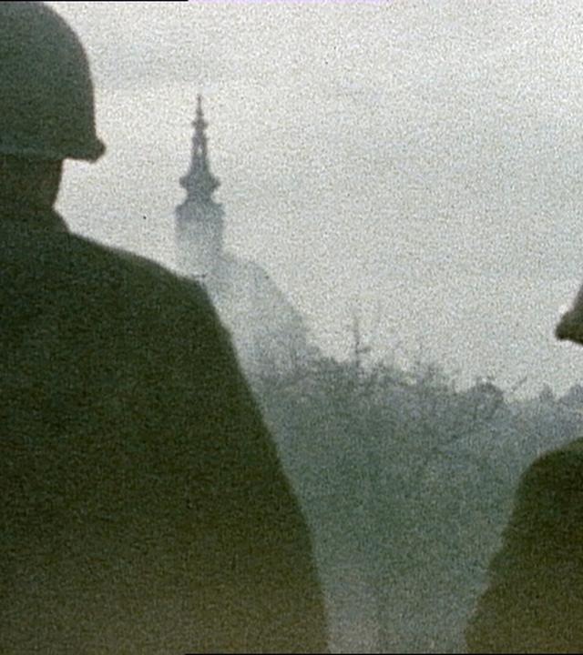 Das Bild zeigt zwei amerikanische Soldaten im Jahr 1945, die in Richtung einer unscharfen Stadtlandschaft blicken. Beide Soldaten tragen helle Stahlhelme und sind von hinten sichtbar. Sie stehen zusammen und blicken auf einen im Hintergrund leicht erkennbaren Turm, der Teil eines Gebäudes ist. Der Himmel über ihnen ist durch einen grauen Schleier verhangen, und die gesamte Szene hat eine melancholische und diffuse Lichtstimmung. Die Umgebung wirkt düster und lässt den Eindruck von Unruhe und Unsicherheit in der Luft entstehen. Es ist klar, dass es sich bei der dargestellten Situation um ein historisches Ereignis zur Zeit des Kriegsendes handelt, wobei die Kulisse vermutlich eine österreichische Stadt im Frühjahr 1945 darstellt.