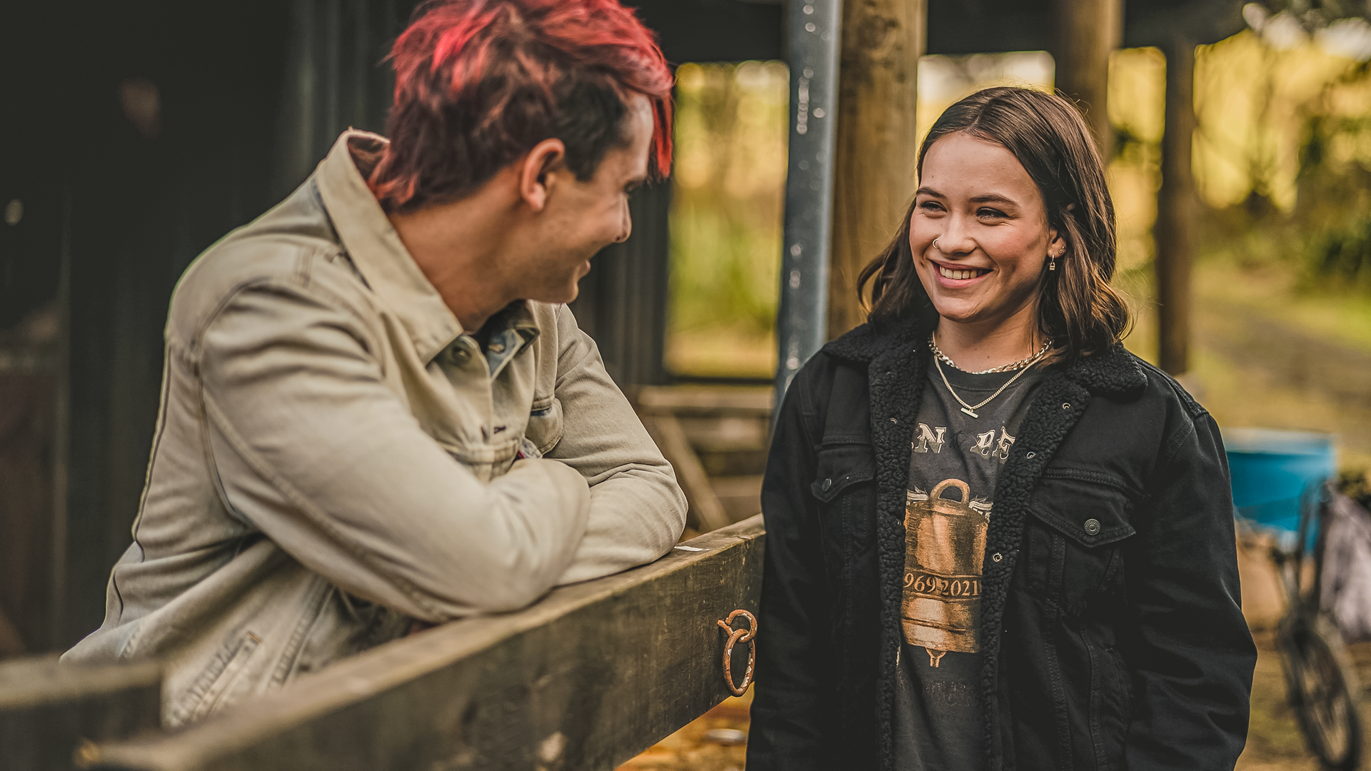 Draußen an einer Pferdebox stehen sich Gabe und Issie gegenüber. Zwischen ihnen ist ein Holzzaun, auf den Gabe sich mit verschränkten Armen stützt. Beide schauen sich mit strahlenden Gesichtern an.