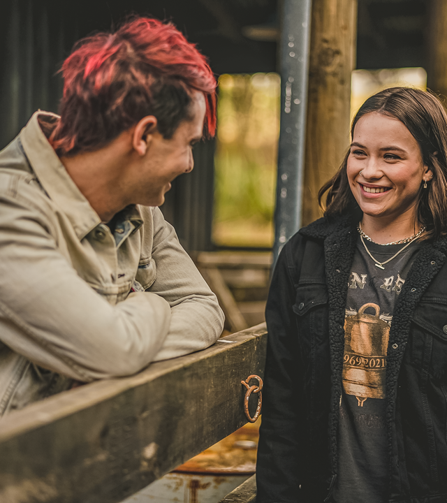 Draußen an einer Pferdebox stehen sich Gabe und Issie gegenüber. Zwischen ihnen ist ein Holzzaun, auf den Gabe sich mit verschränkten Armen stützt. Beide schauen sich mit strahlenden Gesichtern an.