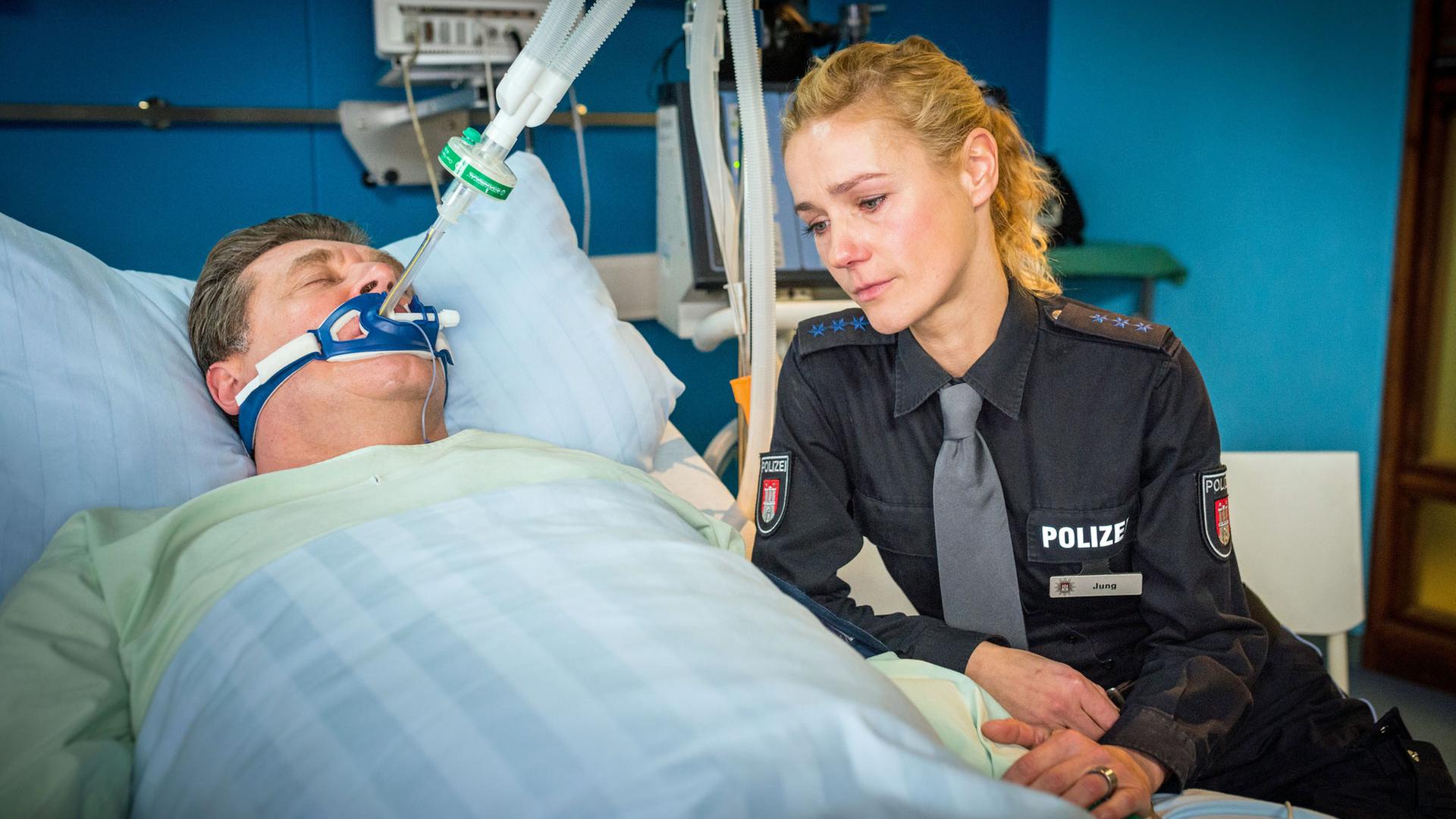 "Notruf Hafenkante - Zwischen Leben und Tod": Hans Moor (Bruno F. Apitz) liegt intubiert in einem Krankenbett im EKH. Er scheint nicht bei Bewusstsein. Die Polizistin Franzi Jung (Rhea Harder-Vennewald) sitzt mit verweinten Augen an seiner Seite und hält seine Hand.