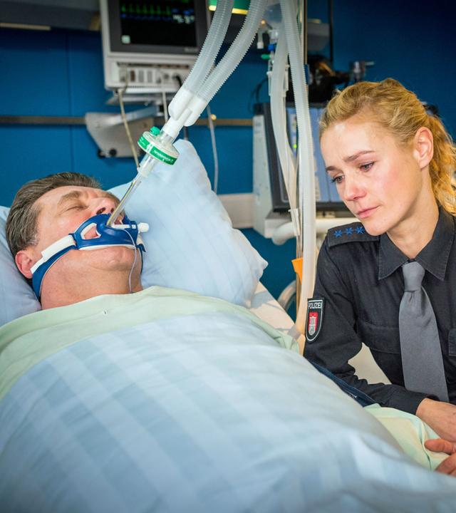 "Notruf Hafenkante - Zwischen Leben und Tod": Hans Moor (Bruno F. Apitz) liegt intubiert in einem Krankenbett im EKH. Er scheint nicht bei Bewusstsein. Die Polizistin Franzi Jung (Rhea Harder-Vennewald) sitzt mit verweinten Augen an seiner Seite und hält seine Hand.