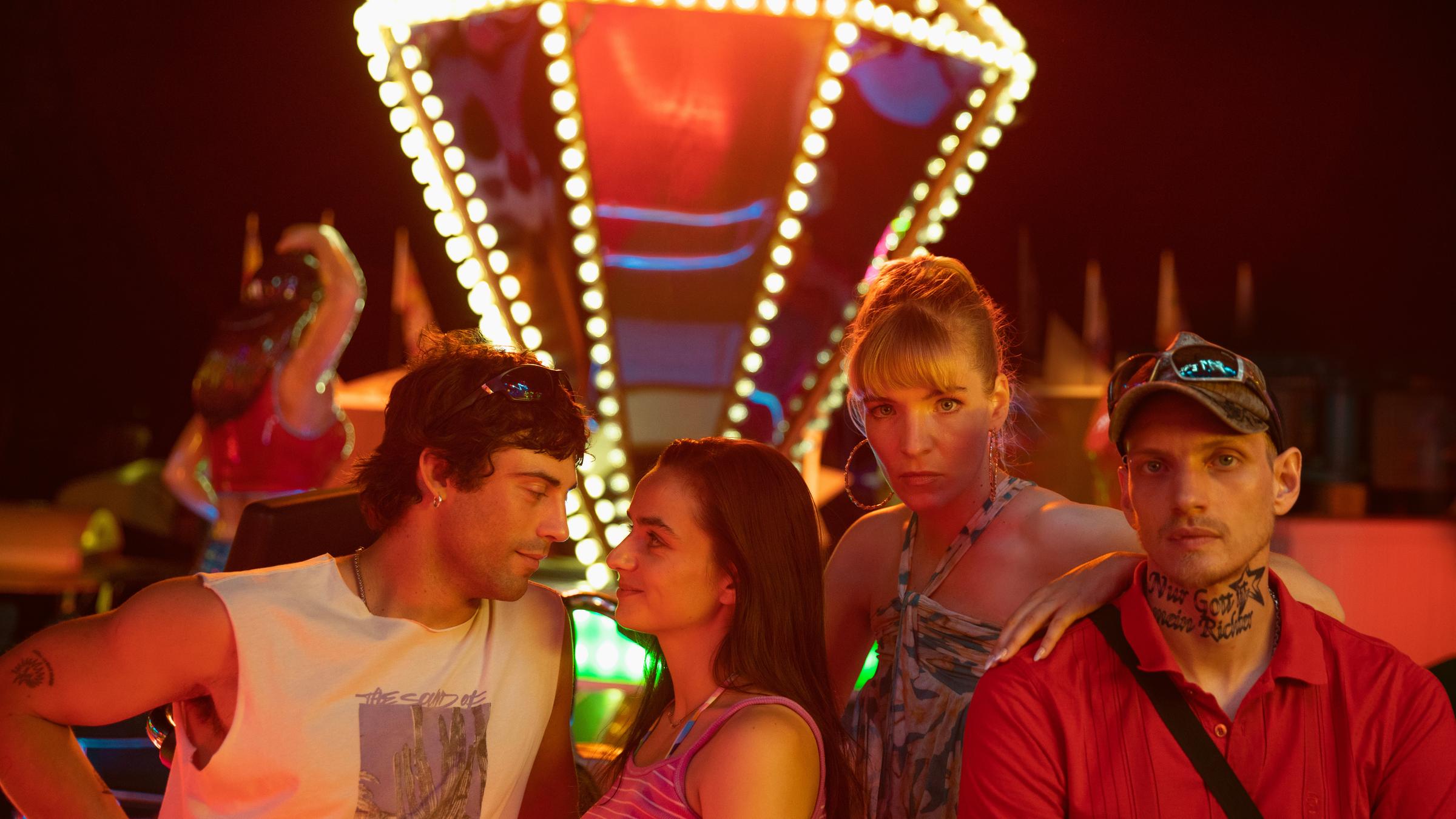 "Zwischen uns die Nacht": Erich (Aaron Altaras) und Marie (Laura Balzer) sehen sich in die Augen während Rebecca (Bea Brocks) und Haro (Paul Boche) in die Kamera sehen. Im Hintergrund leuchtet ein Fahrgeschäft bei Nacht.