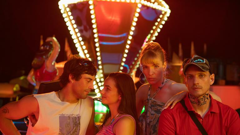 "Zwischen uns die Nacht": Erich (Aaron Altaras) und Marie (Laura Balzer) sehen sich in die Augen während Rebecca (Bea Brocks) und Haro (Paul Boche) in die Kamera sehen. Im Hintergrund leuchtet ein Fahrgeschäft bei Nacht.