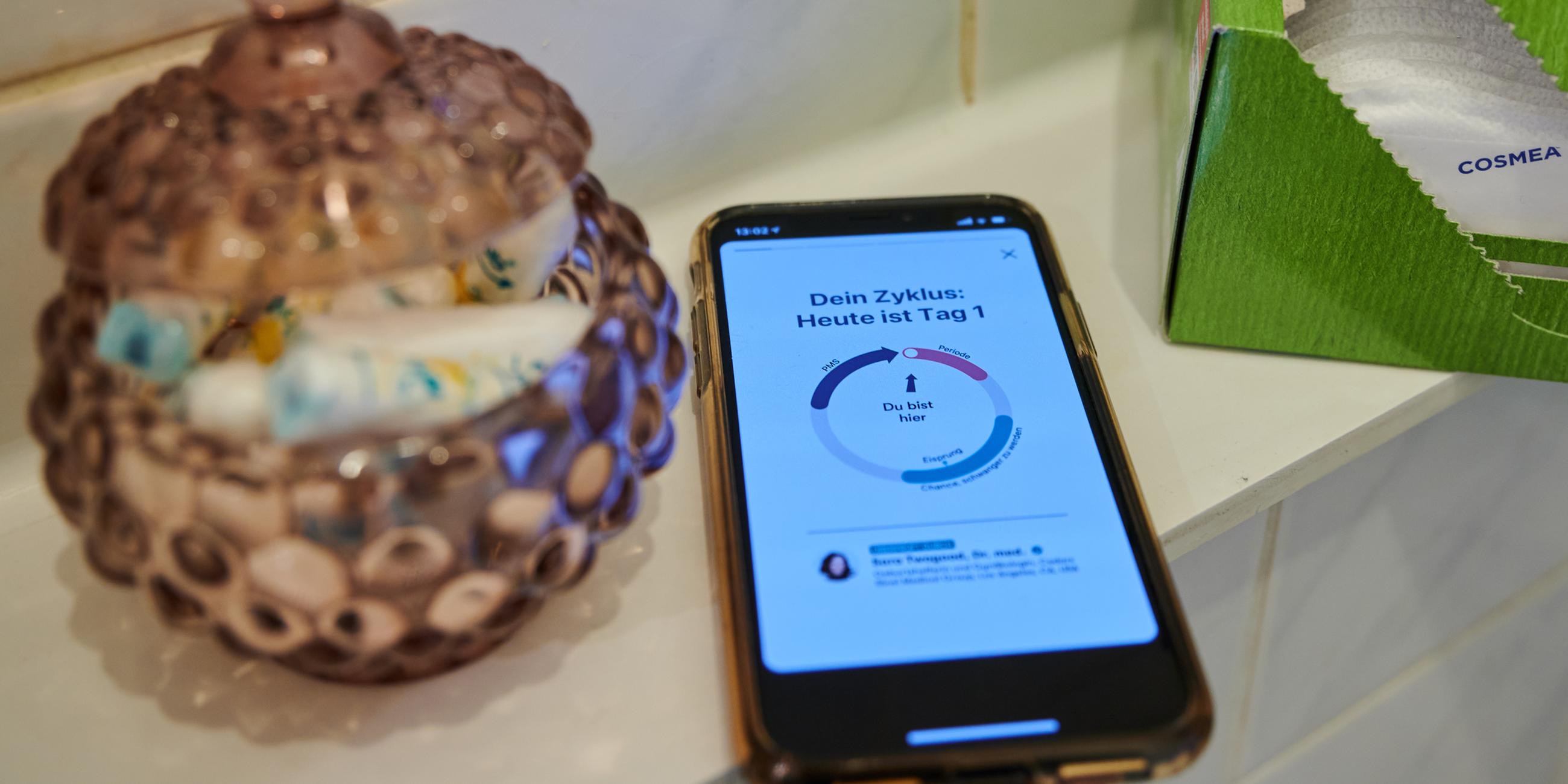 Eine Zyklus-App auf einem Smartphone liegt im Badezimmer neben Slipeinlagen und einem Gefäß mit Tampons. 